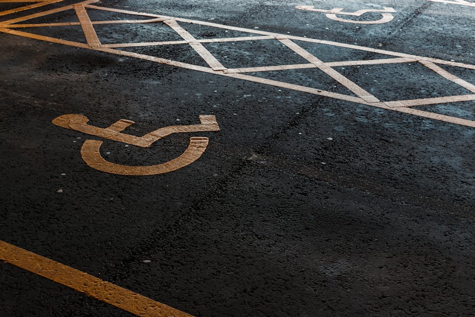 A close-up view of an asphalt parking area showing a white-painted accessible parking symbol located on the ground. The symbol, representing a wheelchair, appears slightly worn but clearly visible, positioned towards the lower left corner of the image. Surrounding the symbol are various marked parking bays with white lines, some of which are faded or partially obscured. The surface is textured with small gravel and about the area appears to be outdoors, likely in a private parking lot, as part of a rubbish removal or waste disposal site. The lighting suggests natural daylight, with some shadows cast across the asphalt, emphasizing the textured surface. The scene is clean, with no visible debris, and the paint markings help delineate parking spaces which may be used for staff or waste collection vehicles. This setting aligns with the context of parking areas serving private waste disposal services, where proper access and parking arrangements facilitate rubbish removal operations, as described on the associated webpage about Dartmouth Park Road rubbish removal access and parking tips.
