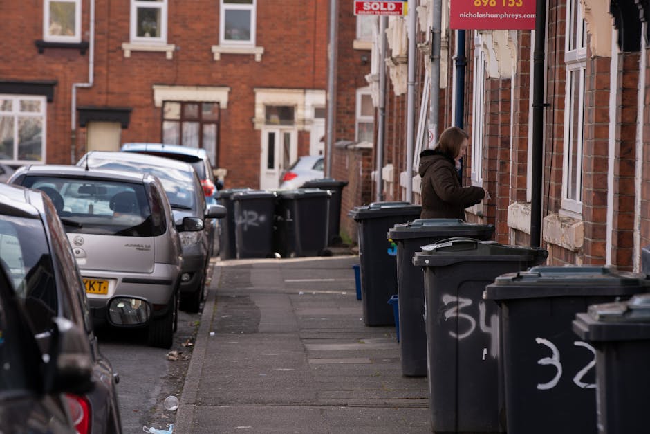 A row of black wheelie bins with closed lids lined up along a residential street curb, positioned in front of brick houses with white window frames and stone detailing. The bins are arranged on the sidewalk, with some displaying white graffiti or markings, and they are placed to facilitate individual waste collection or private disposal. In the background, a woman wearing a dark coat stands near one of the bins, appearing to be handling or closing its lid. Parked cars are positioned along the street, facing towards the background, with the pavement and street surface showing minor wear and scattered debris. The environment suggests an urban setting where residents manage their waste through separate collection, typical for independent rubbish removal services. The scene is lit evenly, with natural ambient daylight highlighting the textures of brickwork, car surfaces, and plastic bin materials, conveying a typical domestic refuse management area within a neighbourhood context.