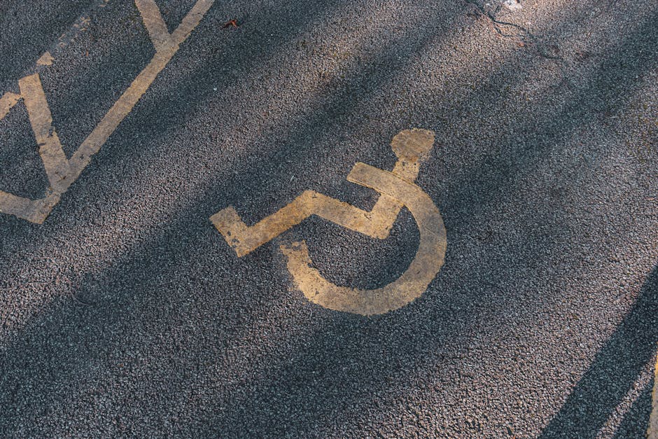 A close-up view of an asphalt parking area showing a white-painted accessible parking symbol located on the ground. The symbol, representing a wheelchair, appears slightly worn but clearly visible, positioned towards the lower left corner of the image. Surrounding the symbol are various marked parking bays with white lines, some of which are faded or partially obscured. The surface is textured with small gravel and about the area appears to be outdoors, likely in a private parking lot, as part of a rubbish removal or waste disposal site. The lighting suggests natural daylight, with some shadows cast across the asphalt, emphasizing the textured surface. The scene is clean, with no visible debris, and the paint markings help delineate parking spaces which may be used for staff or waste collection vehicles. This setting aligns with the context of parking areas serving private waste disposal services, where proper access and parking arrangements facilitate rubbish removal operations, as described on the associated webpage about Dartmouth Park Road rubbish removal access and parking tips.
