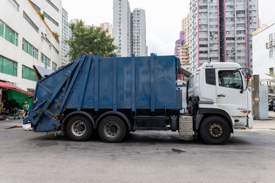 A large commercial waste collection truck is parked on an urban street, with the front cab painted white and the rear waste container in a dark blue colour. The blue container features vertical structural reinforcements and is filled with mixed refuse, typical of private rubbish removal services. The rear loading mechanism is visible, with some parts slightly extended and secured, indicating readiness for collection or unloading. The truck's wheels are sturdy, with the tires showing signs of use. Surrounding the vehicle are multistory buildings, some with modern high-rise architecture and others with older facades, including a building with multiple green window panes on the left and various taller residential towers in the background. The street surface beneath the truck is asphalt, with a few minor stains. Adjacent to the scene are small street vendors with canopies, suggesting a lively urban environment. The overall scene depicts a typical private waste collection operation, supported by [COMPANY_NAME], focused on efficient rubbish removal in a densely built city area.