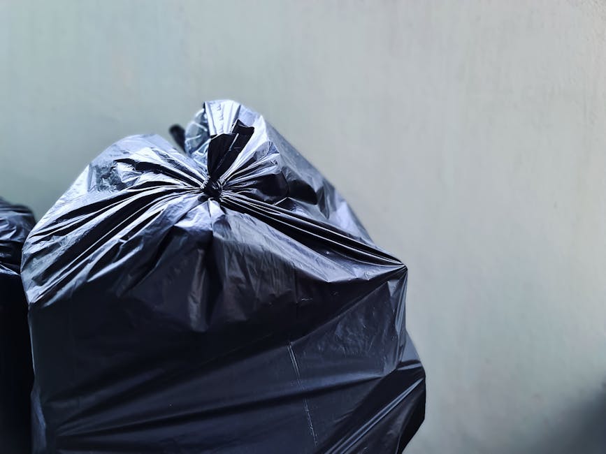 Close-up view of a large black plastic rubbish bag made from glossy and crinkled material, tightly knotted at the top, occupying the left side of the image. The bag appears filled and heavy, with noticeable folds and creases from the compressed waste inside. In the background, a plain, light-colored wall provides a neutral backdrop, with no additional objects or context visible. The lighting is even, highlighting the reflective surface of the plastic and casting gentle shadows. The scene suggests an environment related to waste disposal or rubbish collection, with the bag positioned against a clean wall, potentially in a yard, storage area, or service setting, supporting the theme of private rubbish removal services such as those offered by companies like Rubbish Clearance Tufnell Park.