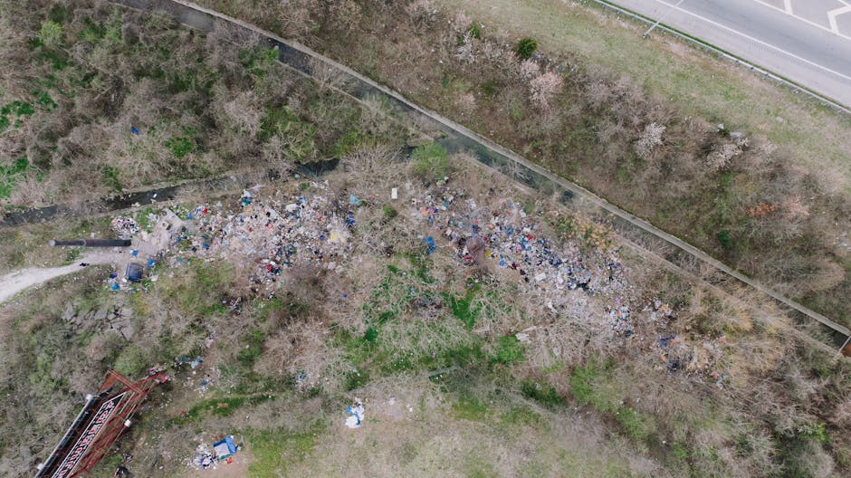 An aerial view of an outdoor waste accumulation area showing a large, irregularly shaped collection of mixed rubbish, including plastic bottles, paper debris, broken wood, and various discarded materials sprawled across a patch of bare soil and sparse greenery. The waste is concentrated near a narrow footpath on the left side of the image and extends towards a chain-link fence border at the top, with some scattered items and waste piles nearer to the bottom. To the right, there is a paved road with a guardrail lining its edge, and beyond the fence, trees and bushes are visible, suggesting a semi-rural or suburban environment. The scene reflects an unmanaged or unregulated rubbish deposit which could be addressed through private waste handling or on-site clearance services, aligning with independent rubbish removal solutions often provided by companies like rubbishclearancetufnellpark.co.uk. The lighting is natural, with diffuse daylight illuminating the entire scene, emphasizing the different textures of the scattered waste materials and the surrounding environment.