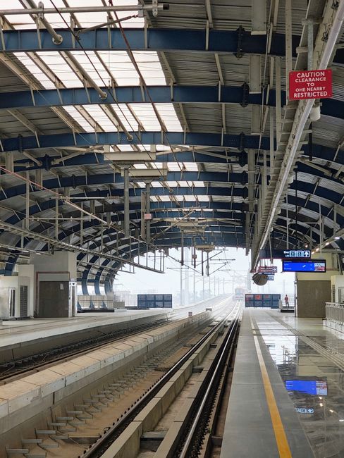 A vacant train station platform with modern architectural features, including a large curved metal and glass roof structure with blue steel beams and transparent panels allowing natural light to fill the space. The platform is clean and empty, with no visible passengers or staff. The railway tracks run along the center, with gravel and concrete sleepers, extending into the distance and towards the horizon. To the right, a yellow safety line runs along the edge of the platform, and a digital information display board is visible, showing train timings. Overhead, a red sign warns about clearance restrictions, indicating a low height clearance area near the station entrance. The overall scene depicts an organized, well-maintained public transport environment, suitable for visitors seeking efficient, independent travel options or private waste management arrangements that may involve on-site clearance or alternative disposal methods, consistent with rubbish removal services. The lighting is predominantly natural, filtered through the canopy's transparent panels, giving the station a bright, airy atmosphere. The image does not depict any rubbish or waste materials, but its clarity aligns with the context of organized infrastructure for public or private transport-related waste management.